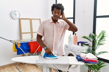 African man with curly hair ironing clothes at home crazy and scared with hands on head, afraid and surprised of shock with open mouth