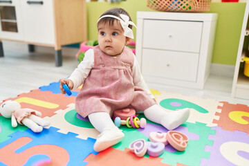 Adorable hispanic baby sitting on floor holding toy at kindergarten