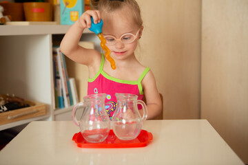 Girl with Down Syndrome learns to pour water from pitcher to pitcher - obrazy, fototapety, plakaty
