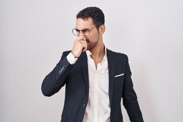 Handsome business hispanic man standing over white background smelling something stinky and disgusting, intolerable smell, holding breath with fingers on nose. bad smell