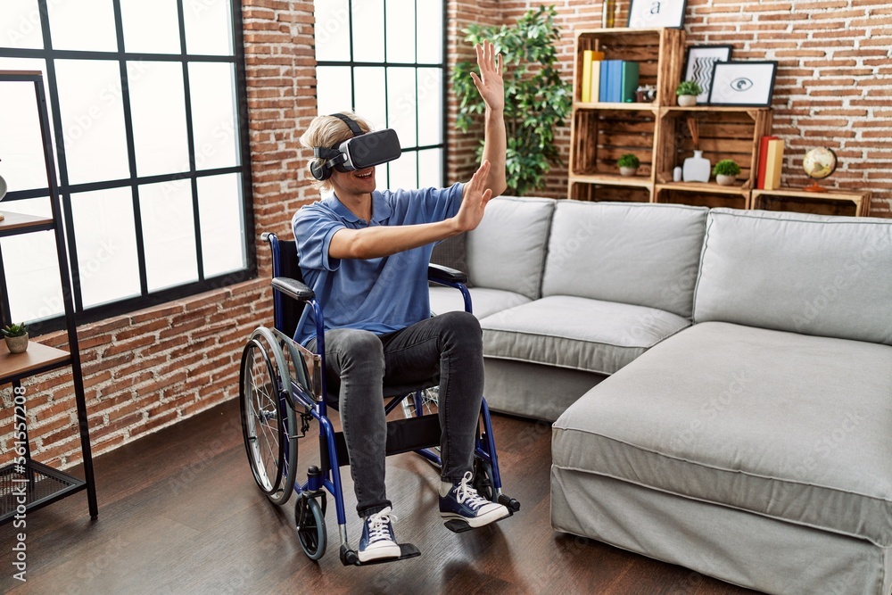 Poster Young blond man playing video game using virtual reality glasses sitting on wheelchair at home