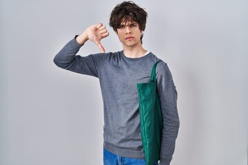 Young man wearing reusable bag with angry face, negative sign showing dislike with thumbs down,...