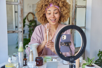 Happy woman wears domestic robe and headband holds cosmetic brush broadcasts live video speaks to followers on her blog gives recommendations sits at table with cosmetics around gives tutorial