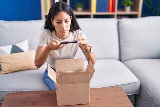 Young beautiful hispanic woman make photo by smartphone unpacking cardboard box at home