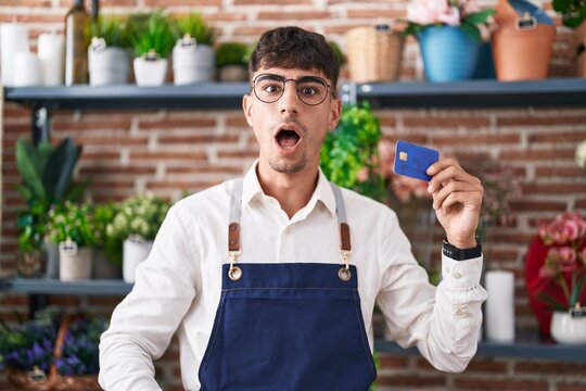 Young hispanic man working at florist shop holding credit card scared and amazed with open mouth for surprise, disbelief face