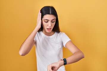 Young beautiful woman standing over yellow background looking at the watch time worried, afraid of...