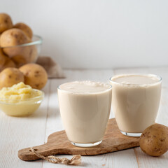 Potato milk in glasses on white wooden background. Pouring vegan milk in glass, with potato puree and potato tubers on background. Copy space. Home made potato milk made from boiled potatoes