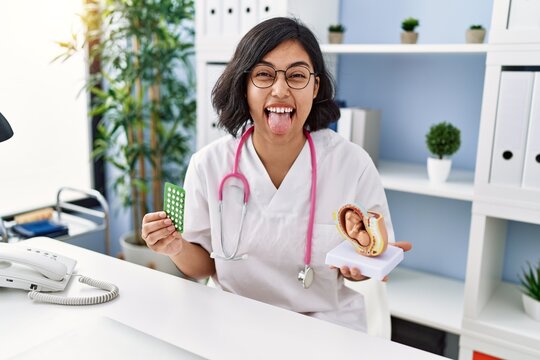 Young Hispanic Doctor Woman Holding Anatomical Model Of Uterus With Fetus And Birth Control Pills Sticking Tongue Out Happy With Funny Expression.