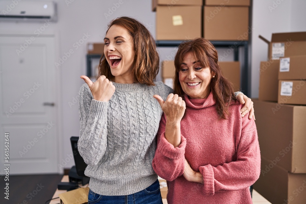 Wall mural Two women working at small business ecommerce pointing thumb up to the side smiling happy with open mouth