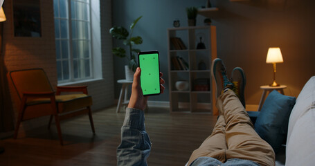 Close up shot of guy lying on couch at night, holding a smartphone with chroma key mock up green screen - technology, connections, communications concept  - obrazy, fototapety, plakaty