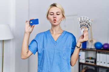 Young physiotherapist woman holding credit card and dollars banknotes making fish face with mouth and squinting eyes, crazy and comical.