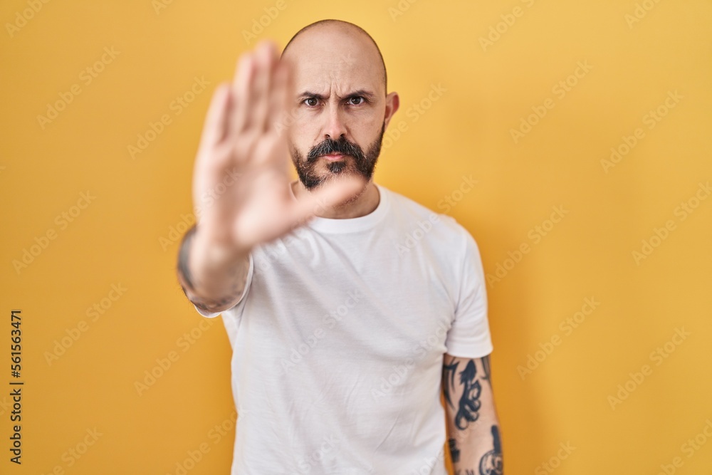 Canvas Prints Young hispanic man with tattoos standing over yellow background doing stop sing with palm of the hand. warning expression with negative and serious gesture on the face.