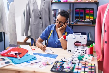 Fototapeta premium African american woman tailor talking on smartphone writing on notebook at tailor shop