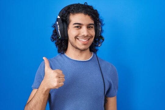 Hispanic man with curly hair listening to music using headphones doing happy thumbs up gesture with hand. approving expression looking at the camera showing success.