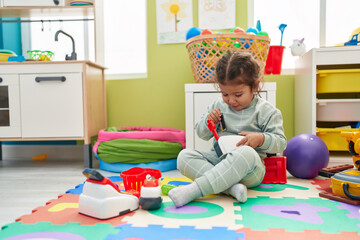 Adorable hispanic toddler playing with supermarket toy sitting on floor at kindergarten