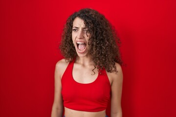 Hispanic woman with curly hair standing over red background angry and mad screaming frustrated and...