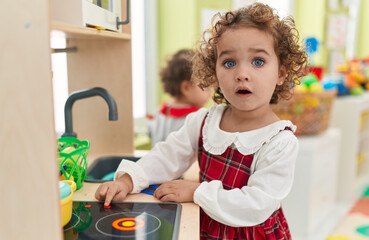 Adorable hispanic toddler playing with play kitchen standing at kindergarten