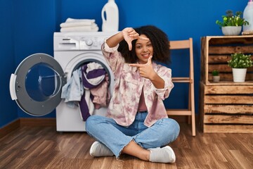 Young african american woman sitting on the floor doing laundry smiling making frame with hands and fingers with happy face. creativity and photography concept.