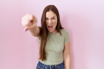 Beautiful brunette woman standing over pink background pointing displeased and frustrated to the...
