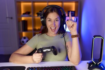 Beautiful brunette woman playing video games wearing headphones pointing finger up with successful idea. exited and happy. number one.