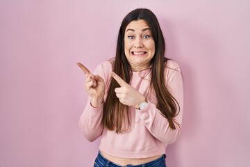 Young brunette woman standing over pink background pointing aside worried and nervous with both hands, concerned and surprised expression