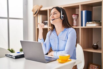Young brunette woman wearing call center agent headset smiling with happy face looking and pointing to the side with thumb up.