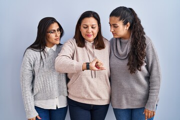 Mother and two daughters standing over blue background checking the time on wrist watch, relaxed and confident