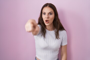 Obraz na płótnie Canvas Young hispanic girl standing over pink background pointing with finger surprised ahead, open mouth amazed expression, something on the front