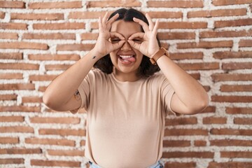 Young hispanic woman standing over bricks wall doing ok gesture like binoculars sticking tongue out, eyes looking through fingers. crazy expression.