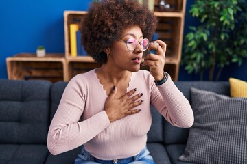 African american woman using inhaler sitting on sofa at home - obrazy, fototapety, plakaty