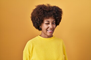 Young african american woman standing over yellow background winking looking at the camera with sexy expression, cheerful and happy face.