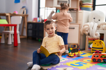 Adorable toddler reading book sitting on floor at kindergarten