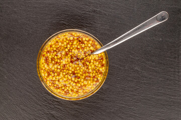 French mustard in a glass saucer with a metal spoon on a slate stone, macro, top view.