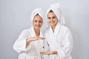 Middle age woman and daughter wearing white bathrobe and towel gesturing with hands showing big and...