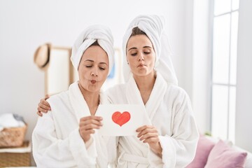 Middle age woman and daughter wearing bath robe holding heart card making fish face with mouth and squinting eyes, crazy and comical.