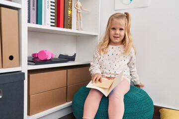 Adorable blonde girl holding book sitting at library school