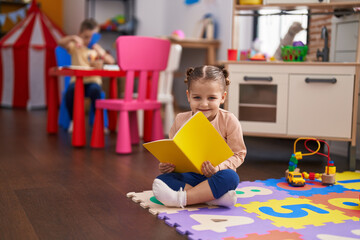 Adorable hispanic girl student sitting on floor reading book at kindergarten