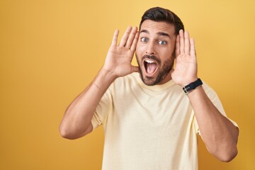 Handsome hispanic man standing over yellow background smiling cheerful playing peek a boo with...