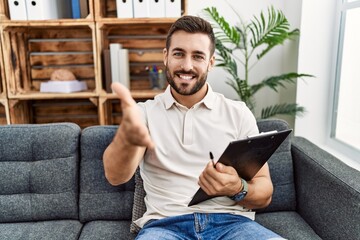 Handsome hispanic man holding clipboard working at psychology clinic smiling friendly offering...