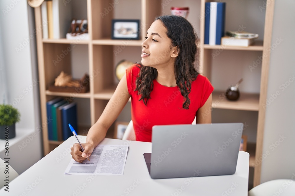 Poster young african american woman using laptop writing on document sitting on table at home