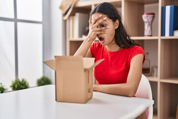 Young brazilian woman looking inside cardboard box peeking in shock covering face and eyes with hand, looking through fingers afraid