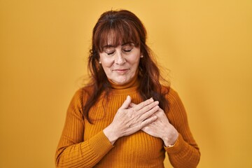 Middle age hispanic woman standing over yellow background smiling with hands on chest with closed...