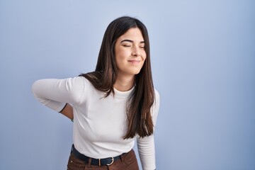 Young brunette woman standing over blue background suffering of backache, touching back with hand, muscular pain