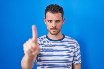 Naklejka premium Young hispanic man standing over blue background pointing with finger up and angry expression, showing no gesture
