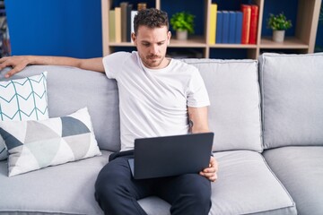 Young hispanic man using laptop sitting on sofa at home
