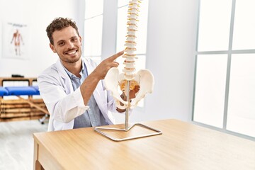 Young hispanic man wearing physiotherapist uniform pointing to anatomical model of vertebral column...