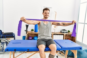 Young hispanic man stretching arm using elastic band sitting on massage board at clinic