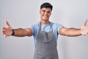 Hispanic young man wearing apron over white background looking at the camera smiling with open arms for hug. cheerful expression embracing happiness.
