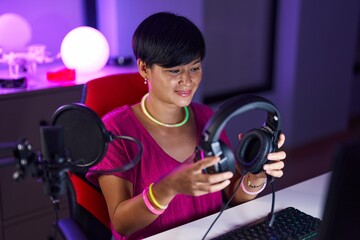 Middle age chinese woman streamer smiling confident holding headphones at gaming room