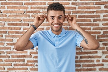 Brazilian young man standing over brick wall smiling pulling ears with fingers, funny gesture....